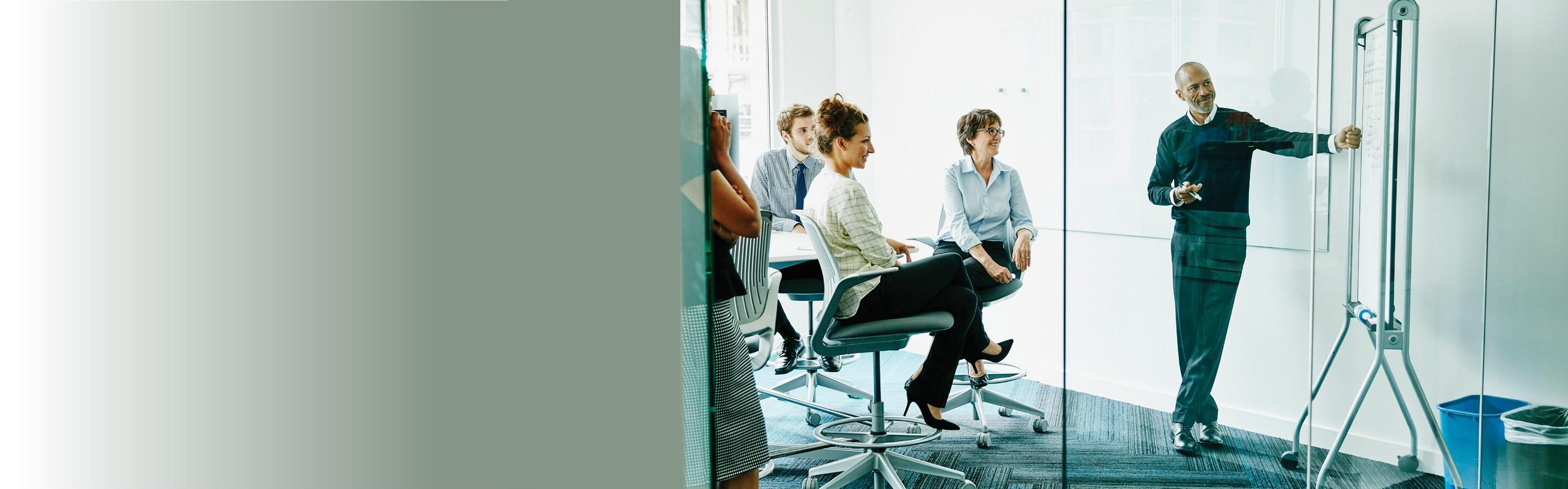 Man presenting in a glass windowed conference room, as coworkers look on smiling and listening